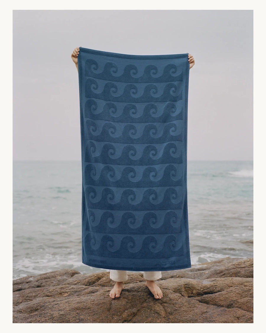 alt=“Individual lounging on Laguna Towel by Laze Res x Journal 1992, on beach with ocean backdrop”